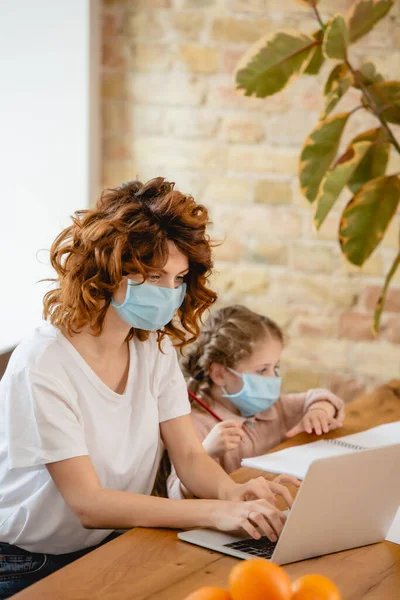 家庭の娘の近くのノートパソコンを使った医療用マスクの中フリーランサーの母親の選択的焦点 — ストック写真