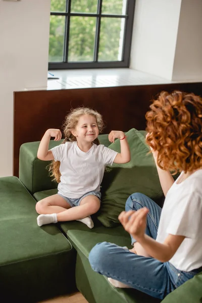 Happy kid looking at curly mother sitting in yoga pose on sofa — Stock Image Happy Kid Looking Curly Mother Sitting Yoga Pose Sofa Stock Image