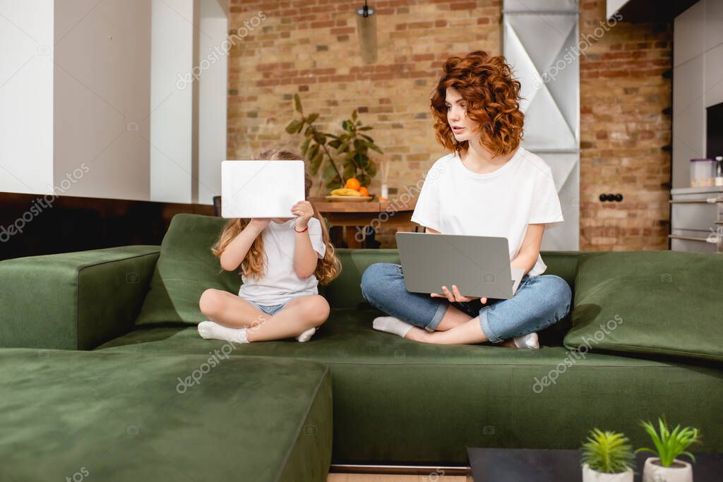 Redhead freelancer using laptop near daughter covering face with digital tablet