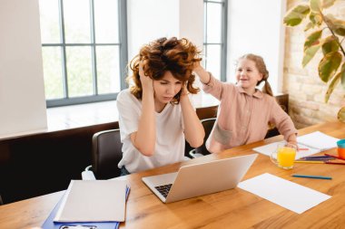 Şirin kız evde laptopun yanında kulaklarını kapatan kıvırcık annesine dokunuyor.