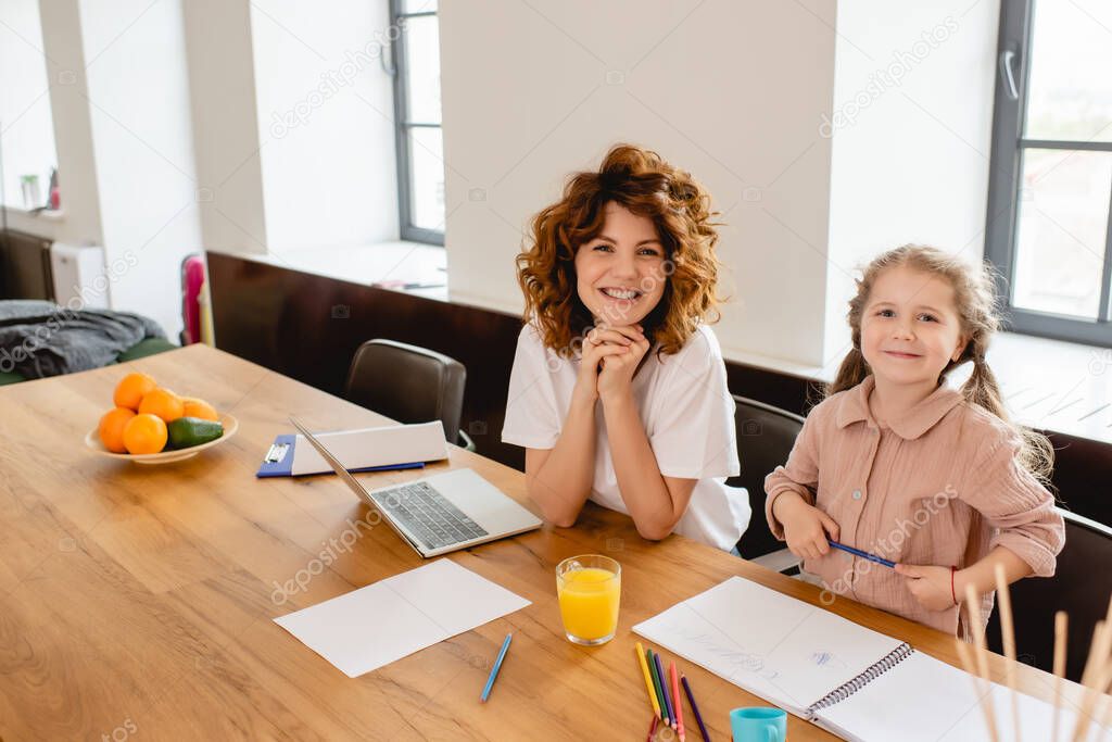 curly freelancer mother smiling near kid drawing near laptop 