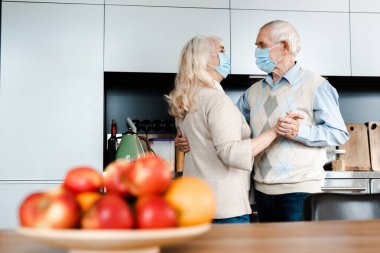Tıbbi maskeli yaşlı çift, izolasyon sırasında mutfakta elmalarla dans ediyor, seçici bir odaklanma.
