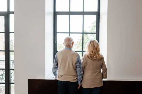 back view of upset elderly couple looking through window at home on quarantine