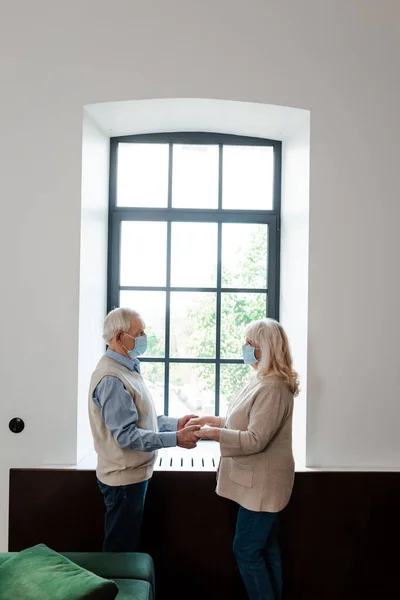 sad elderly couple in medical masks holding hands and standing near window during quarantine