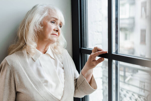 sad elderly woman looking through window during self isolation