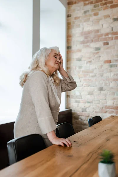 Zmęczona Samotna Seniorka Cierpiąca Ból Głowy Stojąca Przy Stole Podczas — Zdjęcie stockowe