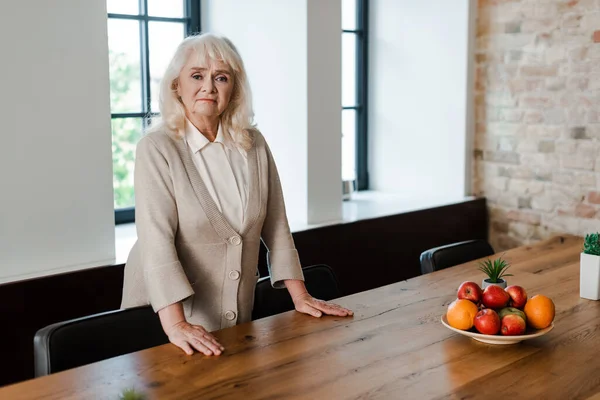 upset senior woman standing at table with fruits during self isolation