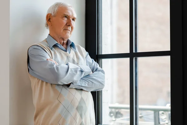 sad lonely man with crossed arms standing near window during self isolation