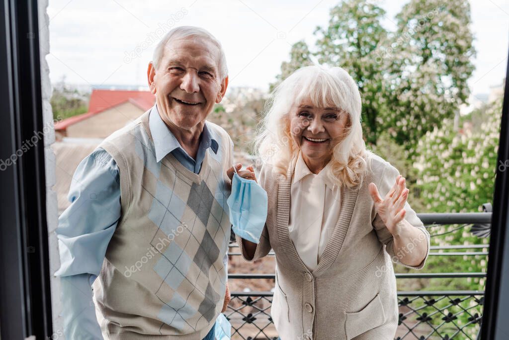 happy elderly couple holding medical masks while standing on balcony after self isolation