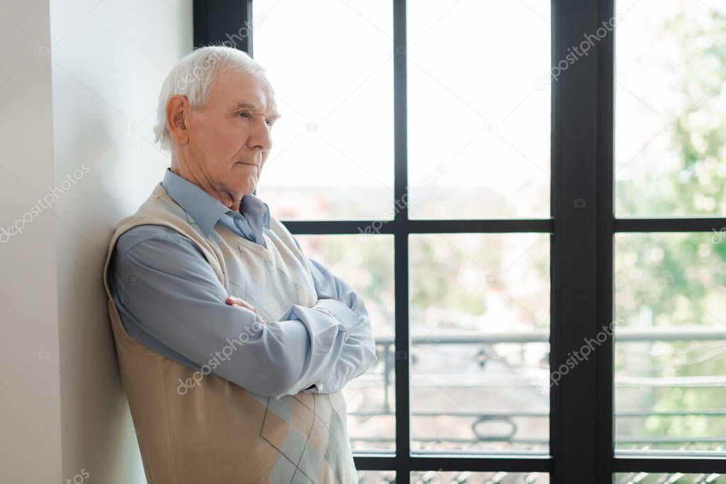 senior lonely man with crossed arms standing near window during self isolation