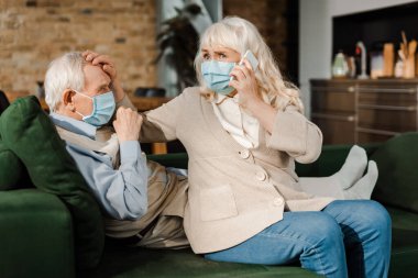 Tıbbi maskeli yaşlı kadın akıllı telefonlu doktoru ararken hasta kocası koronavirüs salgını sırasında kanepede yatıyordu. 