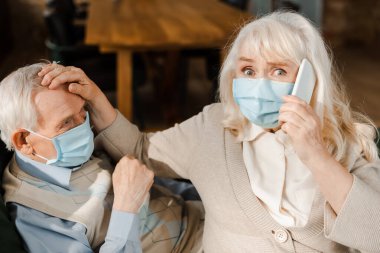 Koronavirüs salgını sırasında hasta kocası kanepede yatarken tıbbi maskeli yaşlı kadın akıllı telefonla doktoru arıyor. 