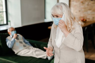 Tıbbi maskeli yaşlı eş akıllı telefonlu doktoru ararken hasta koca koronavirüs salgını sırasında kanepede uzanmış seçici bir şekilde odaklanıyor.