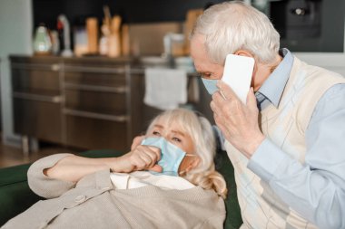 Karantina sırasında hasta bir eş kanepede yatarken tıbbi maskeli yaşlı bir koca akıllı telefonuyla doktoru arıyor.