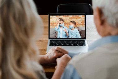 grandparents holding hands and having video chat with grandchildren in medical masks holding thermometer during self isolation clipart