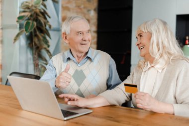 Mutlu yaşlı çift, izolasyon sırasında evde kredi kartı ve laptopla alışveriş yaparken başparmağını gösteriyor.