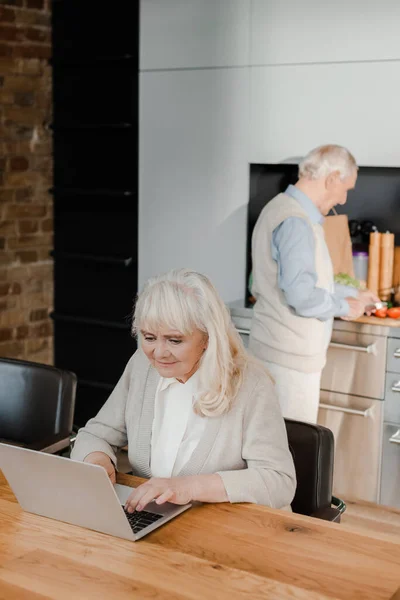 Ηλικιωμένη Σύζυγος Που Χρησιμοποιεί Φορητό Υπολογιστή Ενώ Σύζυγος Μαγειρεύει Στην — Φωτογραφία Αρχείου
