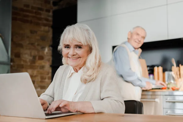 Sorridente Moglie Anziana Utilizzando Computer Portatile Mentre Marito Cucina Cucina — Foto Stock