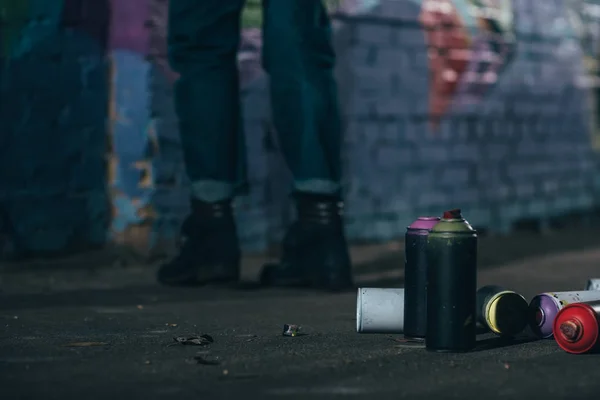 Sección baja del artista callejero pintando graffiti por la noche, latas con pintura en aerosol en primer plano - foto de stock