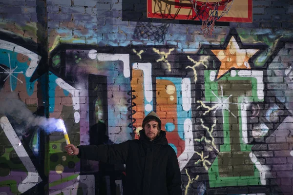 Hombre sosteniendo la bomba de humo y de pie contra la pared con graffiti en la noche - foto de stock