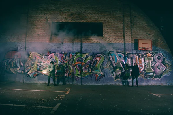 Pessoas segurando bombas de fumaça e de pé contra a parede com graffiti à noite — Fotografia de Stock