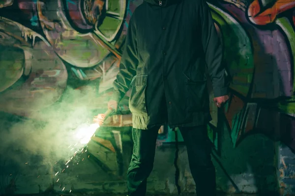 Vue recadrée de l'homme tenant une bombe fumigène et debout contre un mur avec des graffitis la nuit — Photo de stock