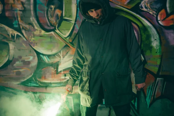 Caucasiano homem segurando bomba de fumaça e de pé contra a parede com graffiti à noite — Fotografia de Stock