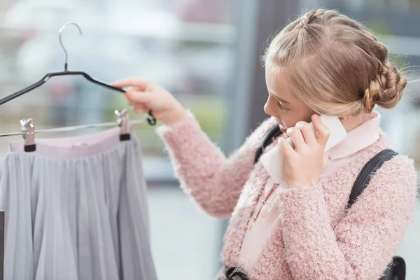 Niño hablando en el teléfono inteligente mientras sostiene tela en la percha en la mano en la tienda - foto de stock