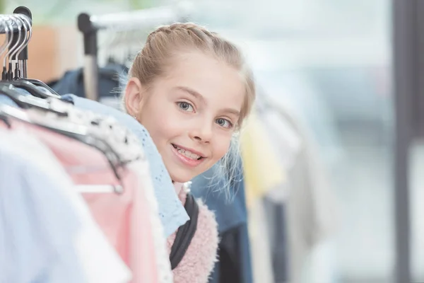 Щаслива дитина дивиться з ряду одягу на вішалці в магазині — стокове фото