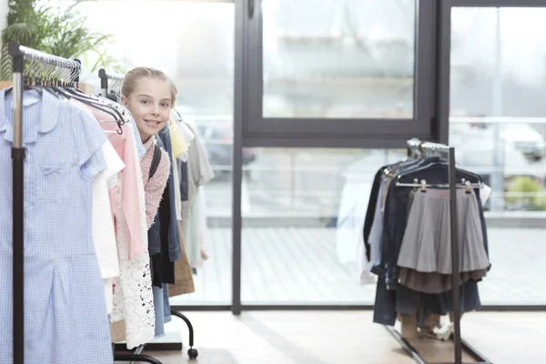 Niño sonriente mirando desde la fila de ropa en la percha en la tienda - foto de stock