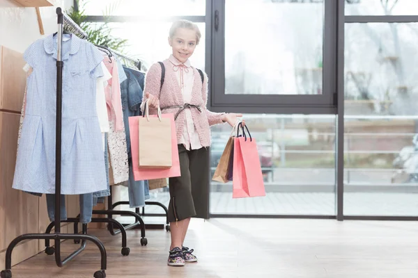 Smiling kid holding paper shoping bags in hands while standing near hanger at shop — Stock Photo