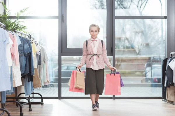 Улыбающийся ребенок, держащий в руках бумажные сумки для покупок, окруженный вешалками в магазине — стоковое фото