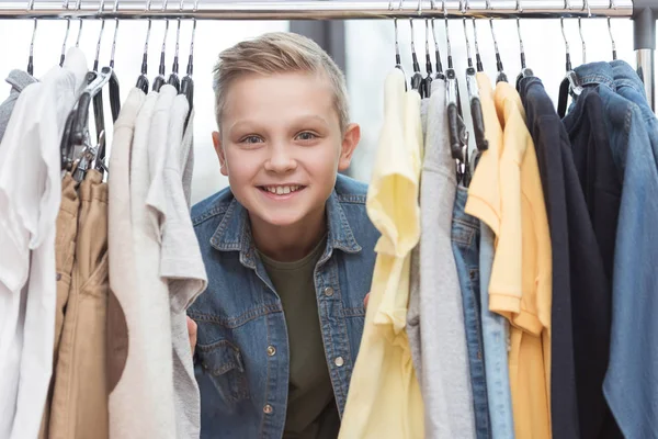 Lächelnder Junge blickt in die Kamera, umgeben von Kleidern auf Kleiderbügel im Geschäft — Stockfoto