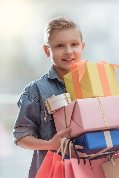 Niño sonriente sosteniendo cajas con bolsas de papel de colores en las manos en el interior de la tienda - foto de stock