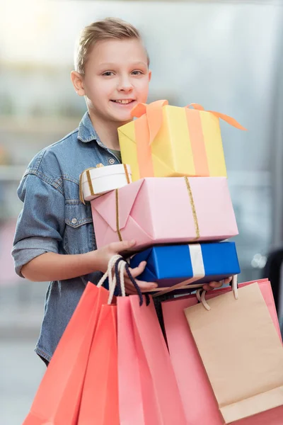 Niño sonriente sosteniendo cajas con bolsas de papel en las manos en el interior de la tienda - foto de stock