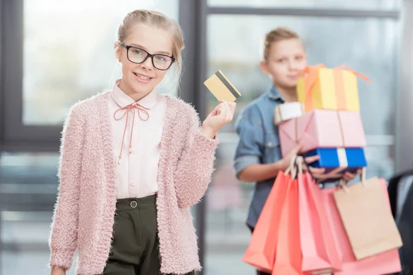 Garoto em óculos segurando cartão de crédito na mão com menino segurando caixas no fundo — Fotografia de Stock