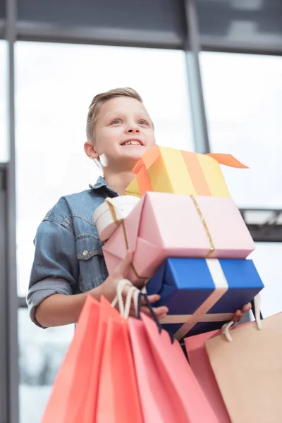 Menino feliz segurando caixas coloridas com sacos de papel nas mãos na loja interior — Fotografia de Stock