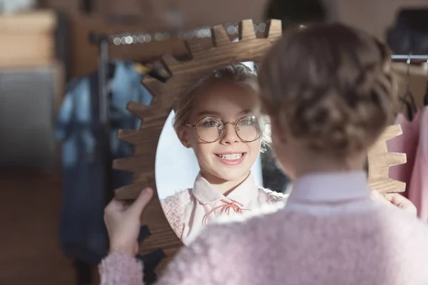 Happy child in glasses looking at mirror in her hands at store interior — Stock Photo