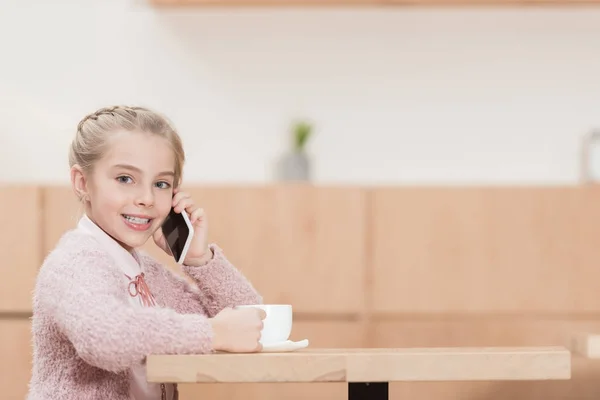 Lächelndes Kind sitzt mit Tasse in der Hand und benutzt Smartphone, während es in die Kamera schaut — Stockfoto