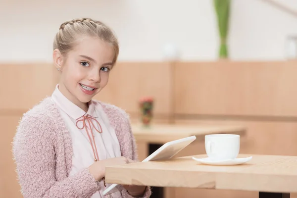 Усміхнена дитина з цифровим планшетом дивиться на камеру, сидячи за столом — стокове фото