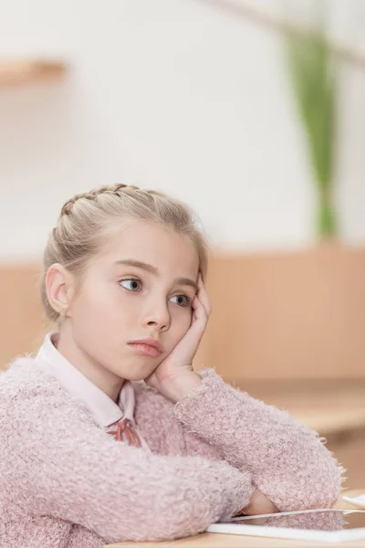 Niño aburrido sentado en la mesa con la tableta digital y mirando hacia otro lado - foto de stock