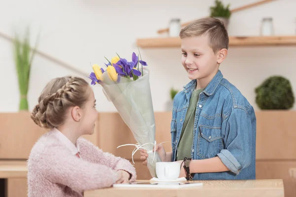 Menino sorrindo apresentando flores para menina sentada à mesa namorada — Fotografia de Stock