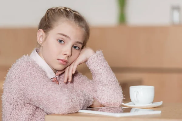 Enfant ennuyé assis à table avec tablette numérique et regardant la caméra — Photo de stock