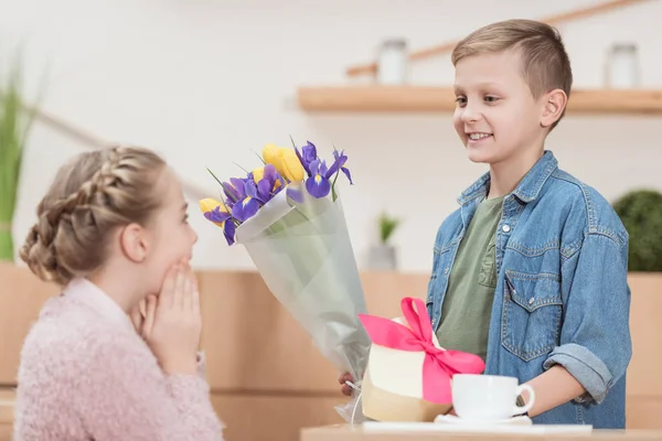 Smiling boy presenting bouquet and box with ribbon to surprised girlfriend at cafe — Stock Photo