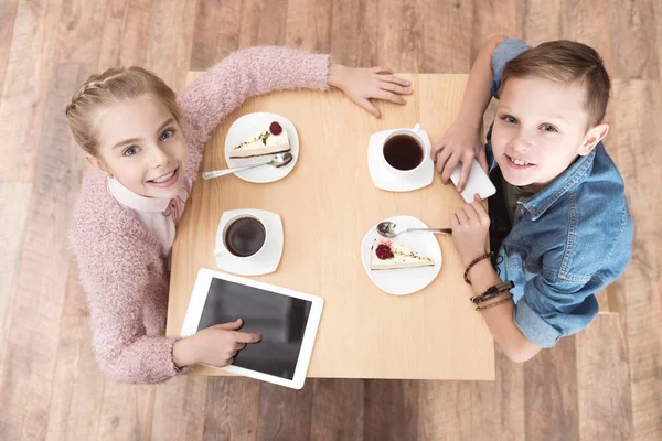 Діти дивляться на камеру, сидячи за столом з гаджетами на поверхні в кафе — стокове фото