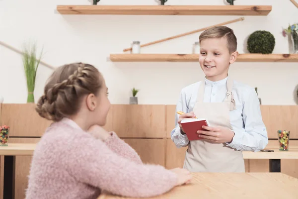 Petit serveur souriant prendre des notes dans le bloc-notes contre l'enfant au café — Photo de stock