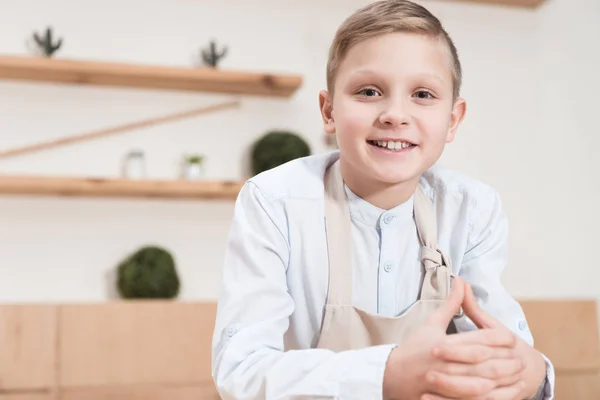 Lächelnder Junge in Schürze, der in die Kamera blickt, während er sich im Café auf den Tisch lehnt — Stockfoto