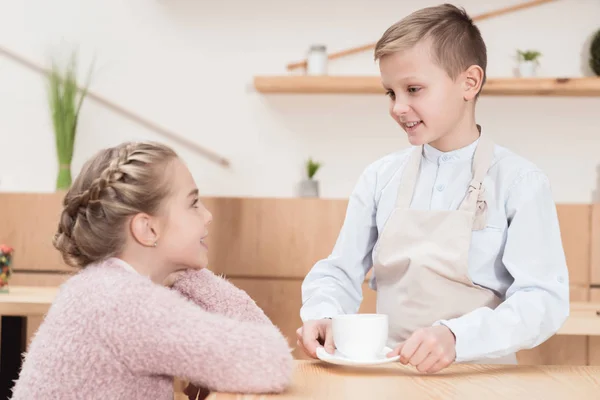 Pequeño camarero con taza en las manos mirando a un niño sentado en la mesa en la cafetería - foto de stock