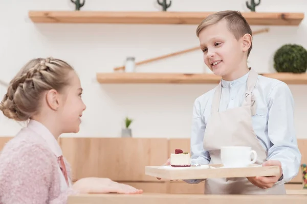 Pequeño camarero sosteniendo bandeja de madera en las manos con postre contra el niño sentado en la mesa en la cafetería - foto de stock