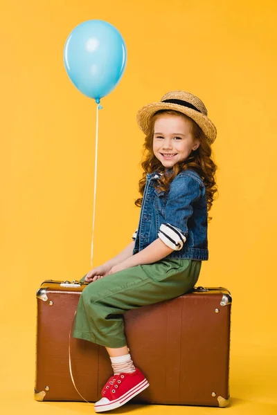 Vista lateral del niño con globo sentado en la maleta aislado en amarillo - foto de stock